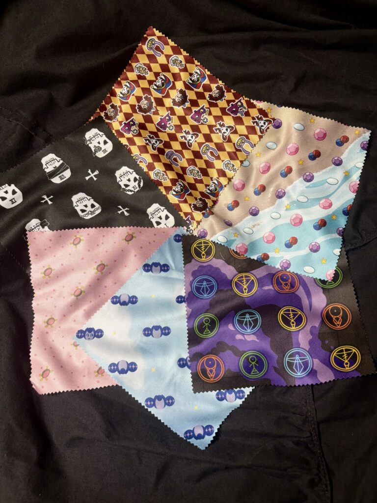 A photo of six decorative glasses cloths, in designs referencing The Amazing Digital Circus, Steven Universe, The Owl House, Knights of Guinevere, Infinity Train, and The Locked Tomb.